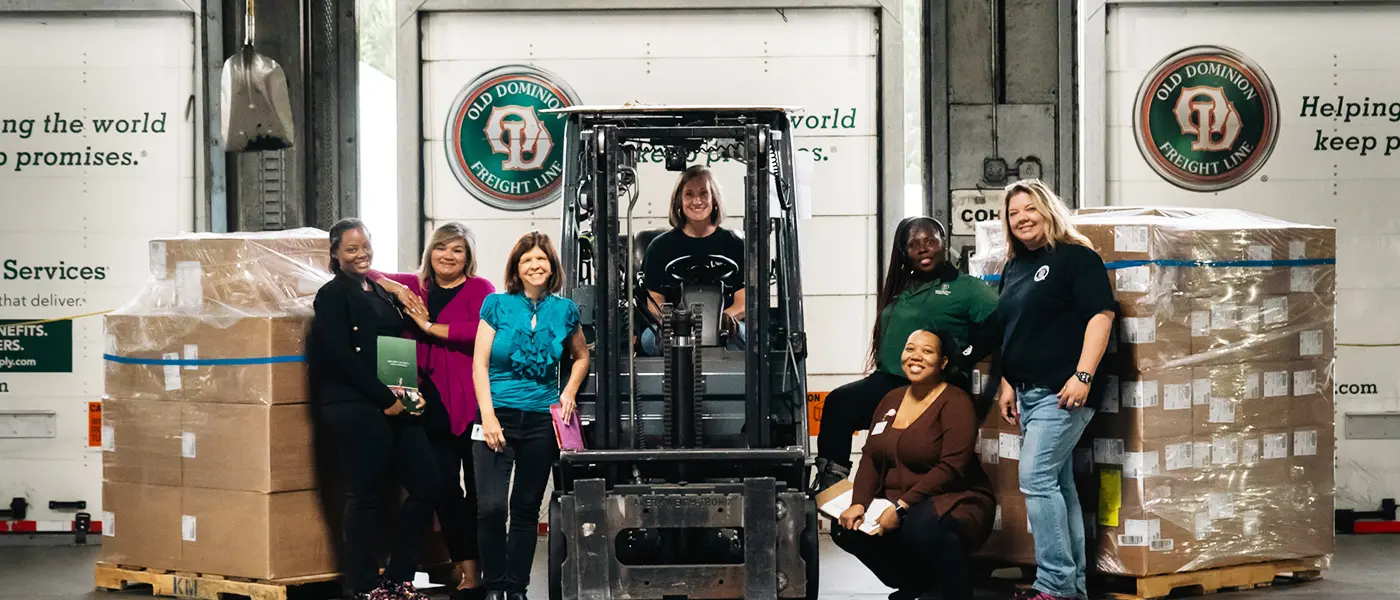A group of women in transportation posing with a forklift in front of Old Dominion Freight Line trucks and pallets.